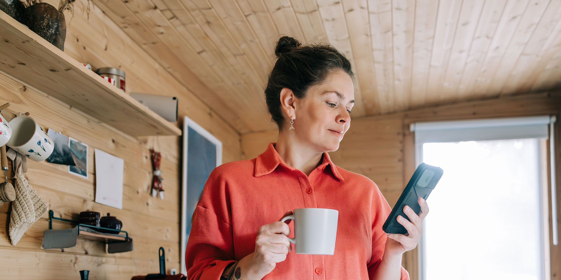 A person looking at their phone and holding a cup of coffee while standing in the kitchen of their wood cabin.