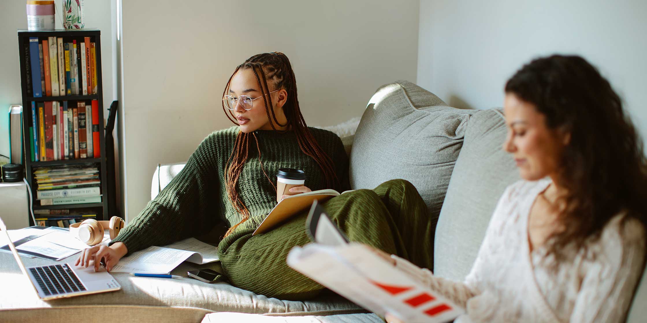 Two people sit on a sofa in their living room. One reads a book; the other holds coffee and scrolls on a laptop.