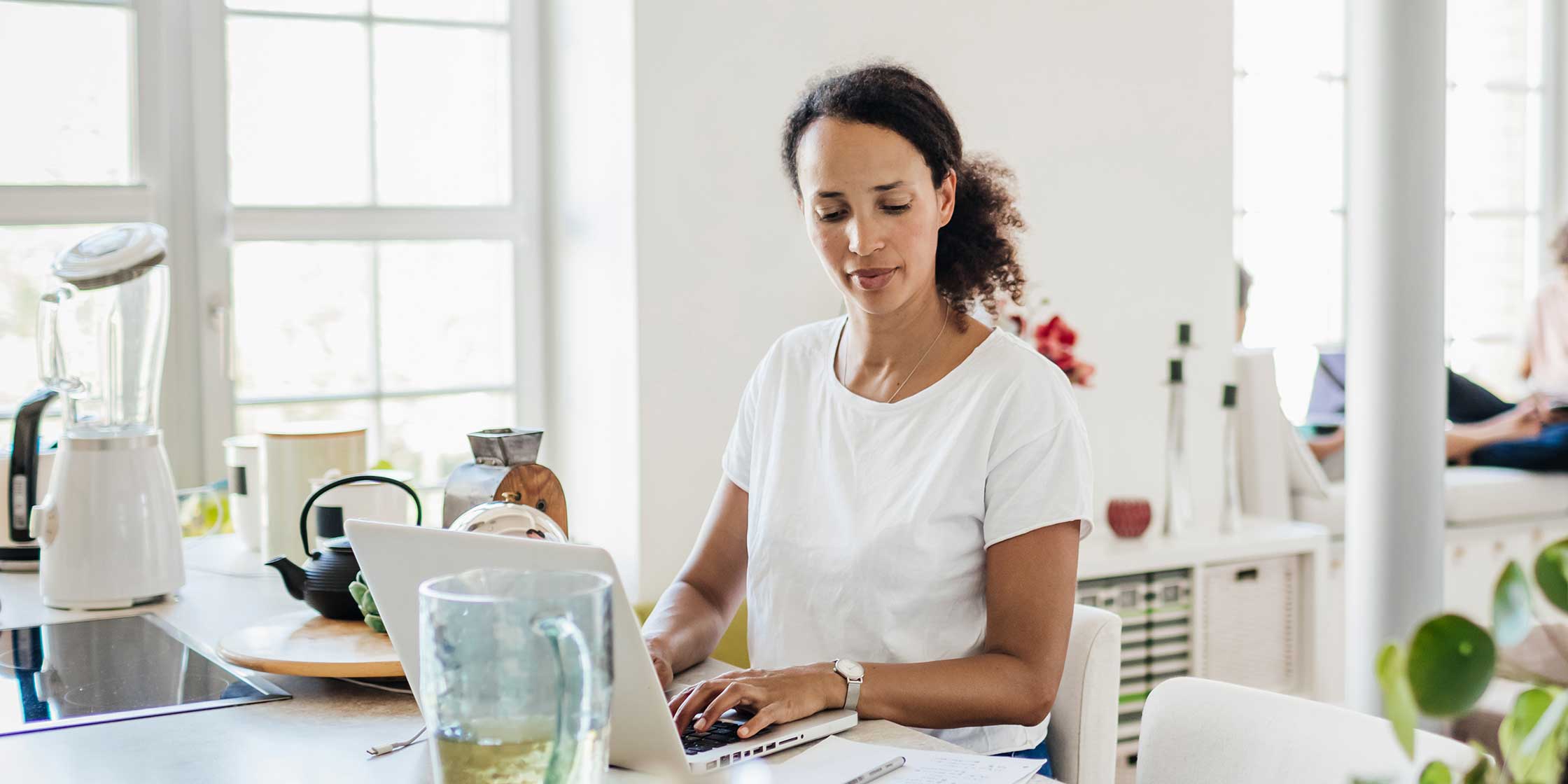 A mom sits at her kitchen counter, using her laptop while her kids read in the background.