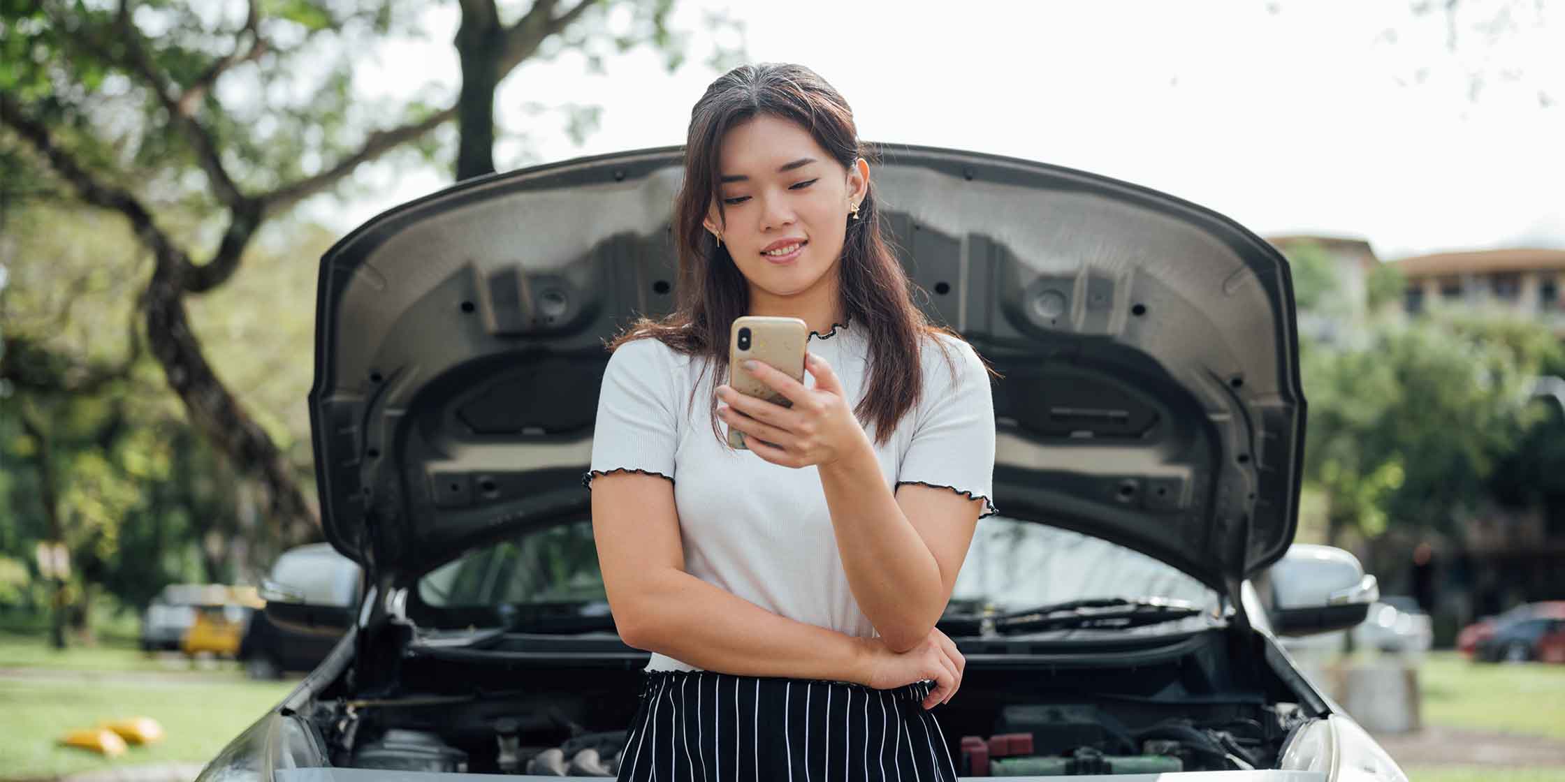A person standing in front of their open car hood and looking at their phone while waiting for roadside assistance.