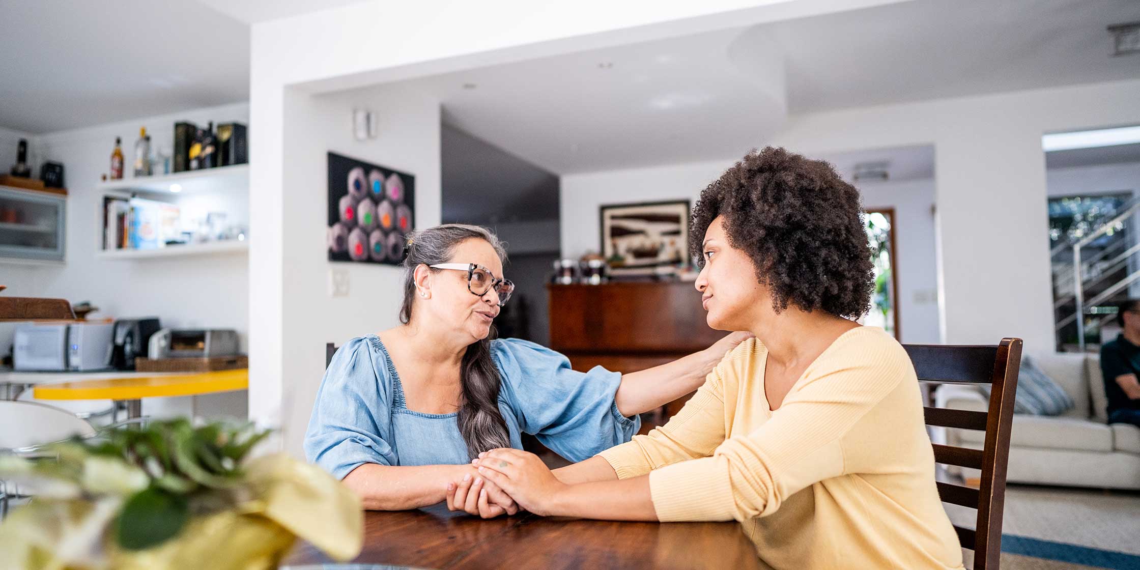 Two people sit at a kitchen table in deep conversation, clasping hands.