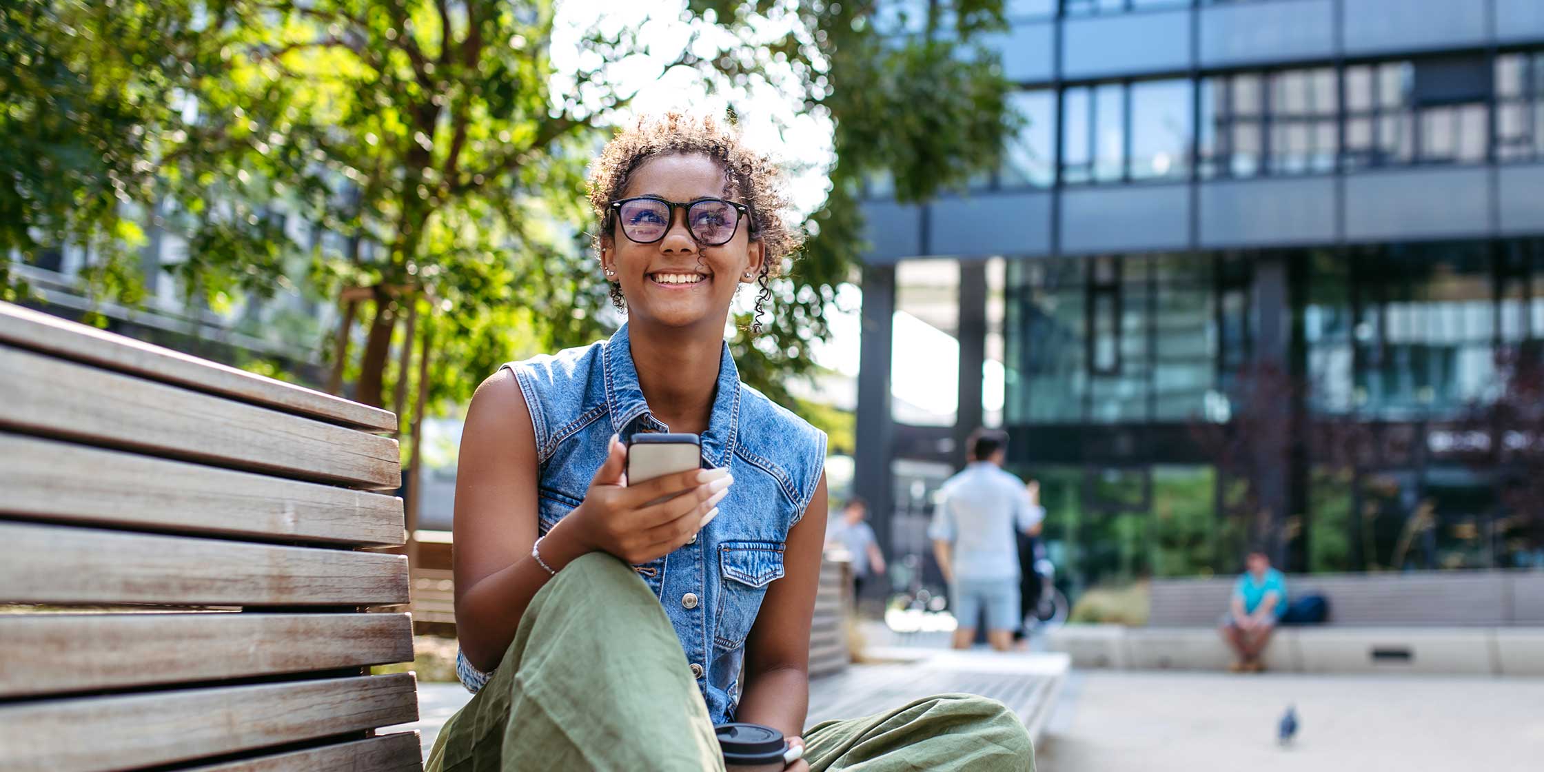 A college student sits on a bench with their phone in hand, reading about building credit while in school.