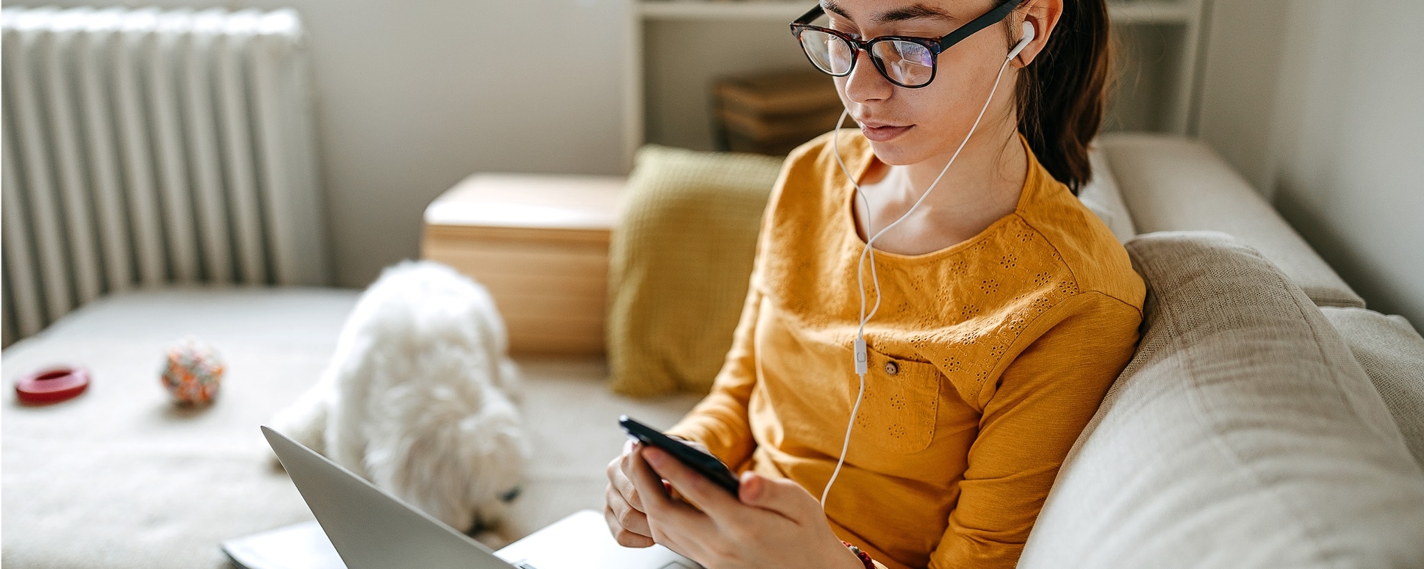 A college student wearing earbuds sits on her couch and checks her phone.
