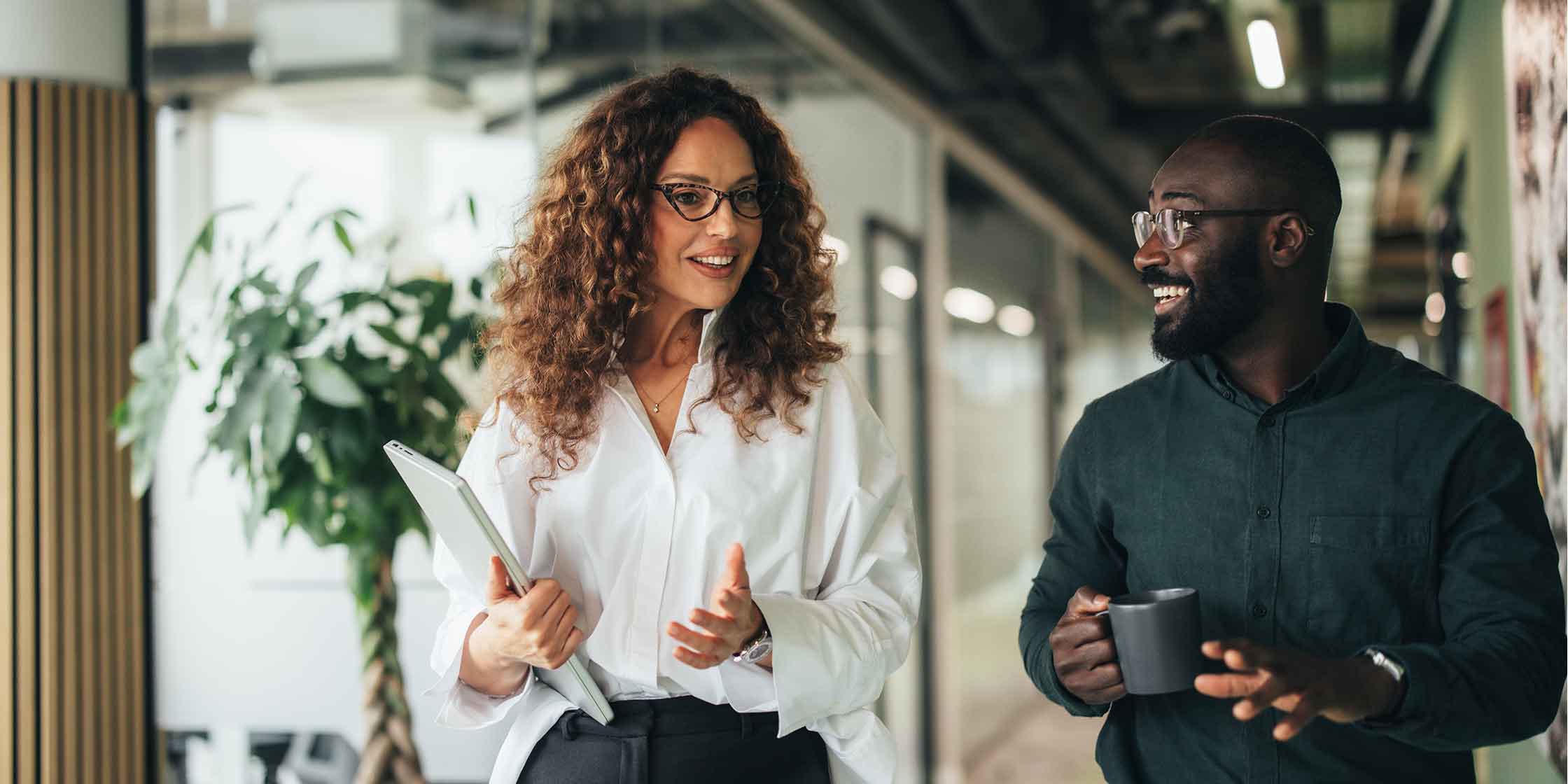 Two smiling people dressed in business casual chatting while walking in an office.