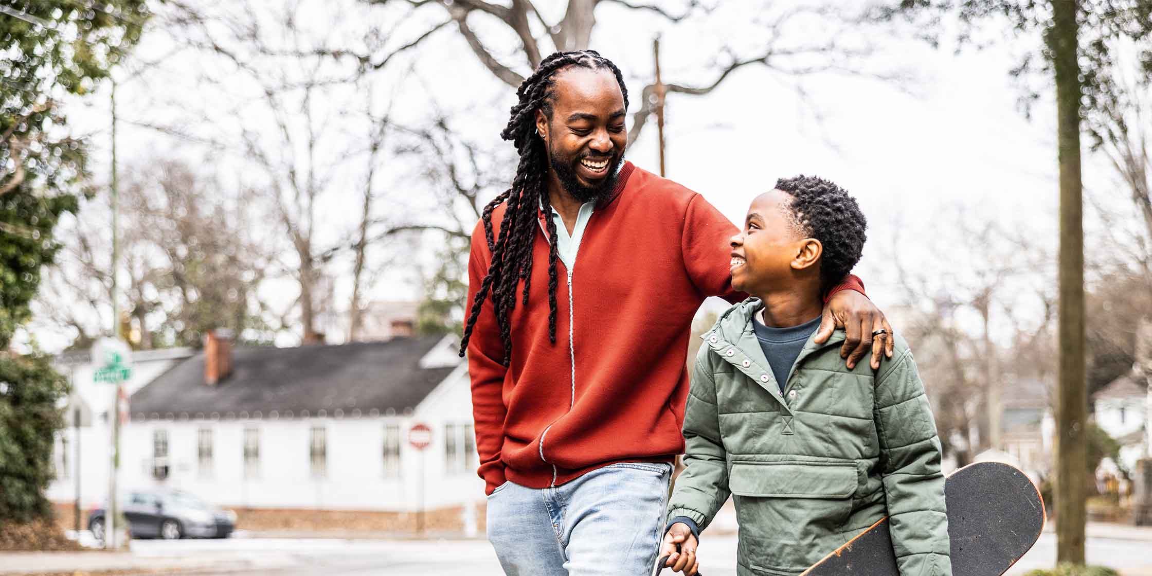 A smiling father puts his arm around his son as they walk outside.