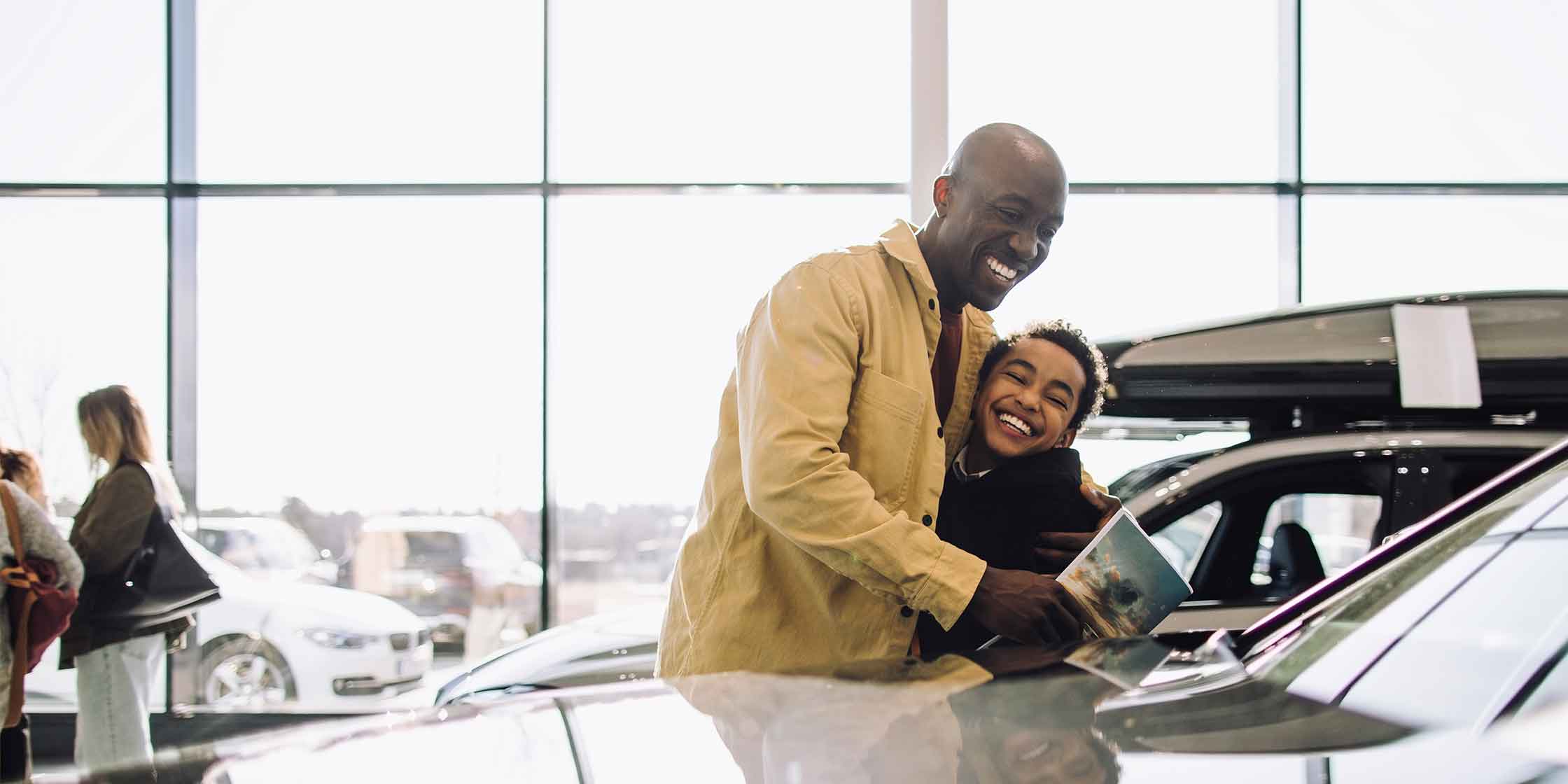 Man and child excitedly hug while looking at a new car in a dealership.