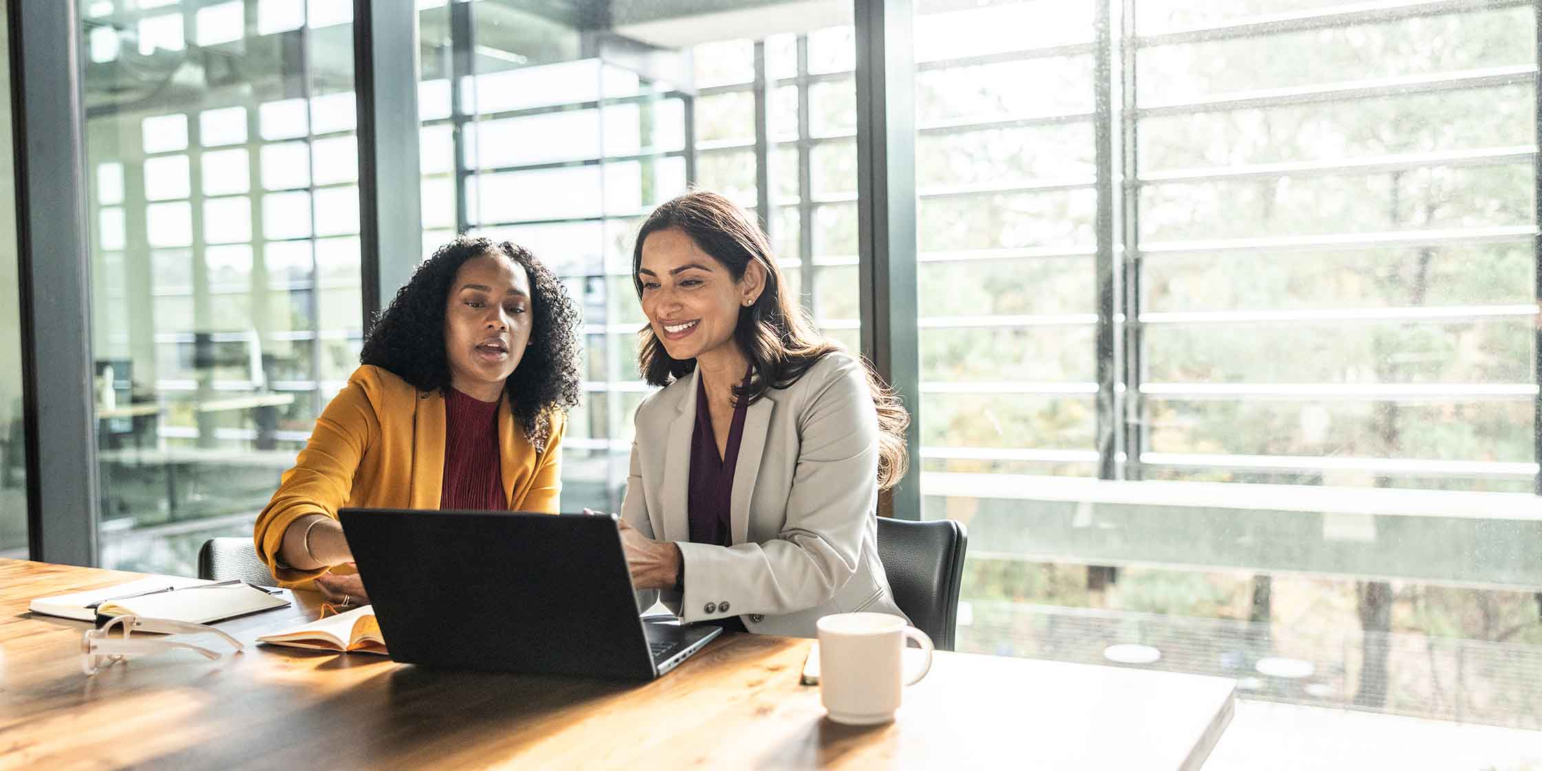 Two women look at a laptop in an office conference room.