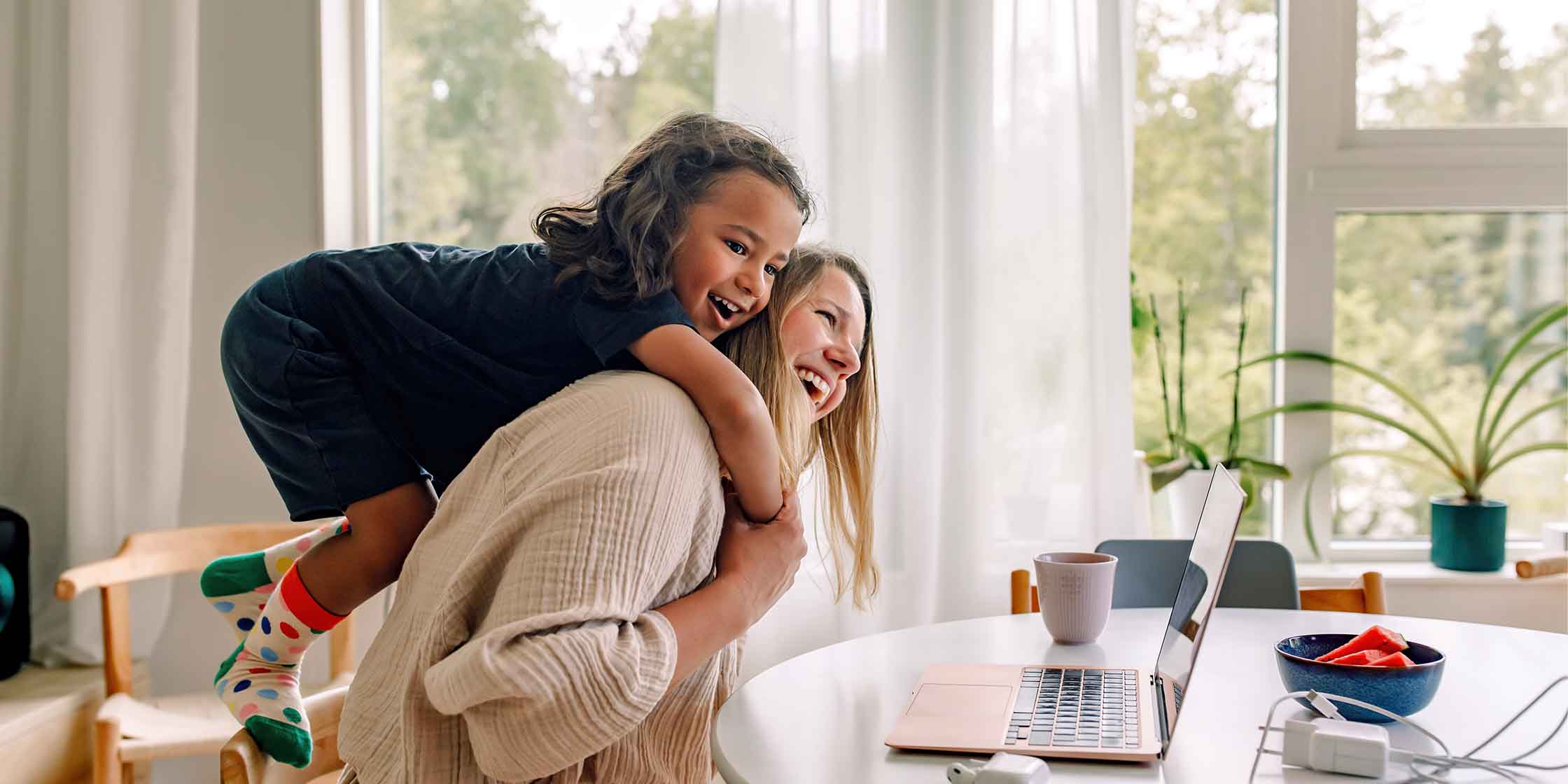 A child hugs their parent around the neck from behind while they both laugh.