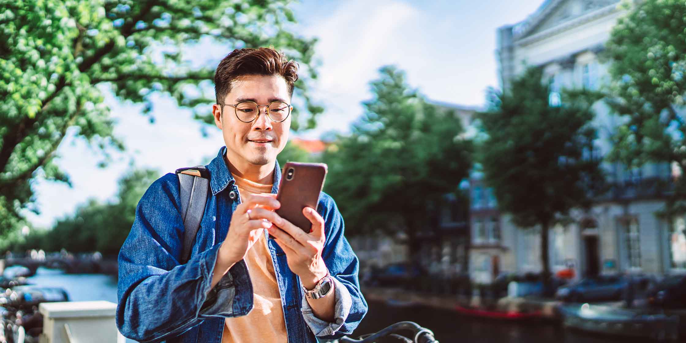 A person wearing glasses, a denim shirt and a backpack walks down a city street while using a smartphone.