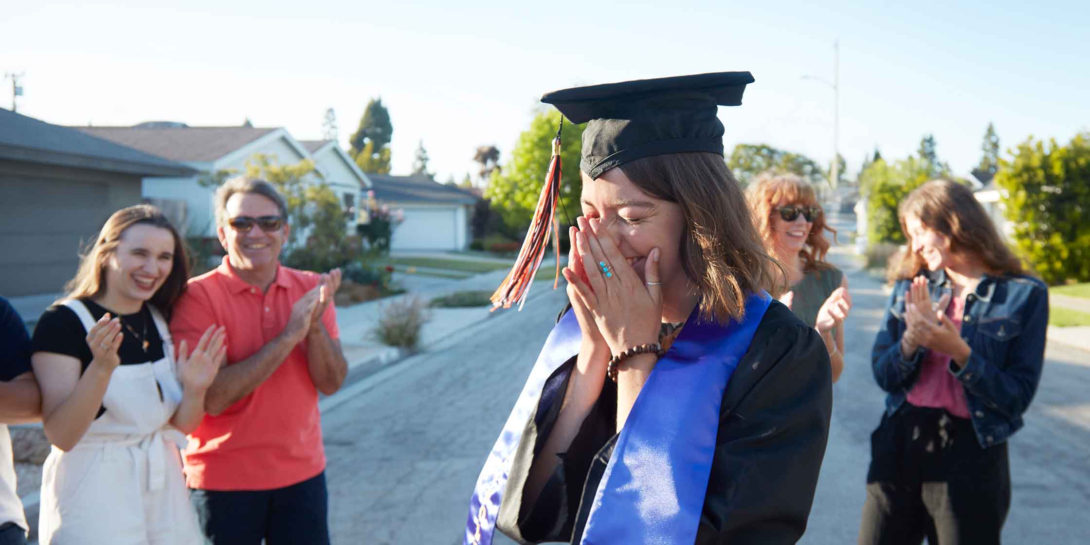 A student in graduation garb stands in the middle of a neighborhood street smiling as friends and family clap and smile around her.