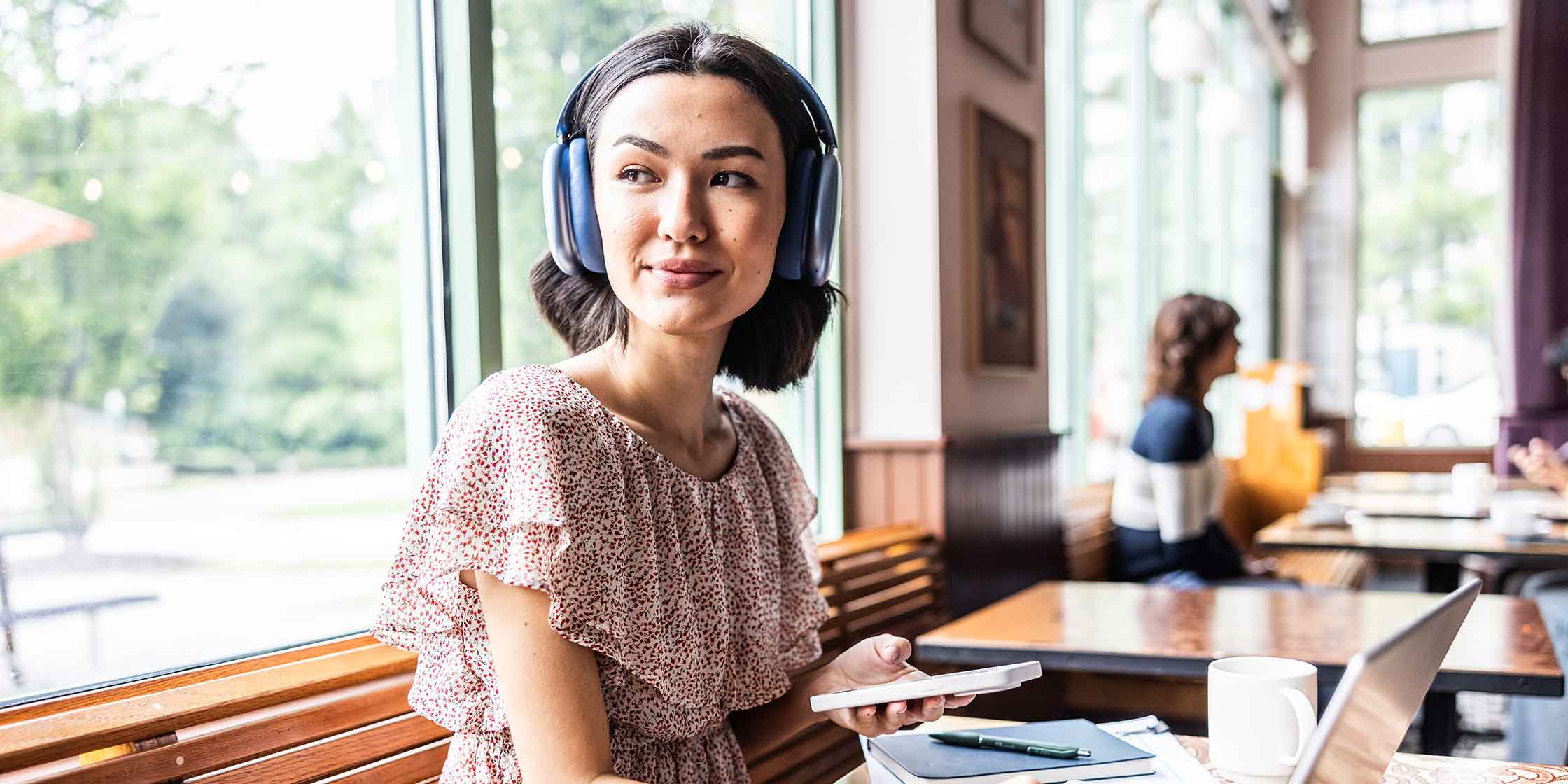 A young person wearing headphones with a phone and laptop sits at a table in a cafe.