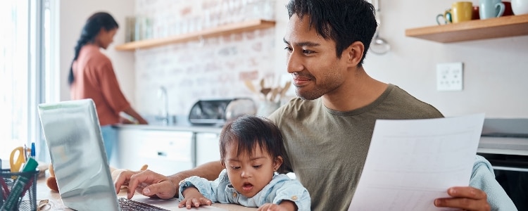 A parent with their baby on their lap sits at a kitchen table and uses a laptop.