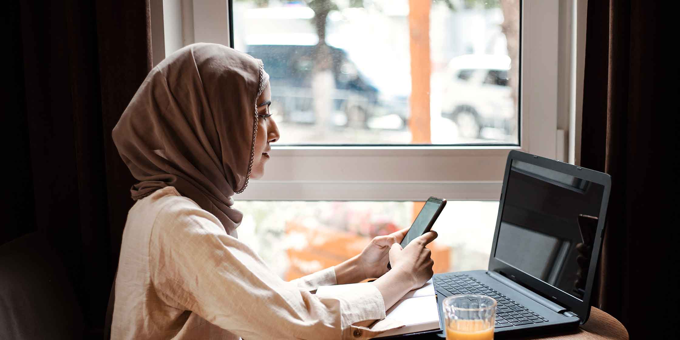 A woman wearing a hijab sits in front of a window, using her cell phone and laptop.