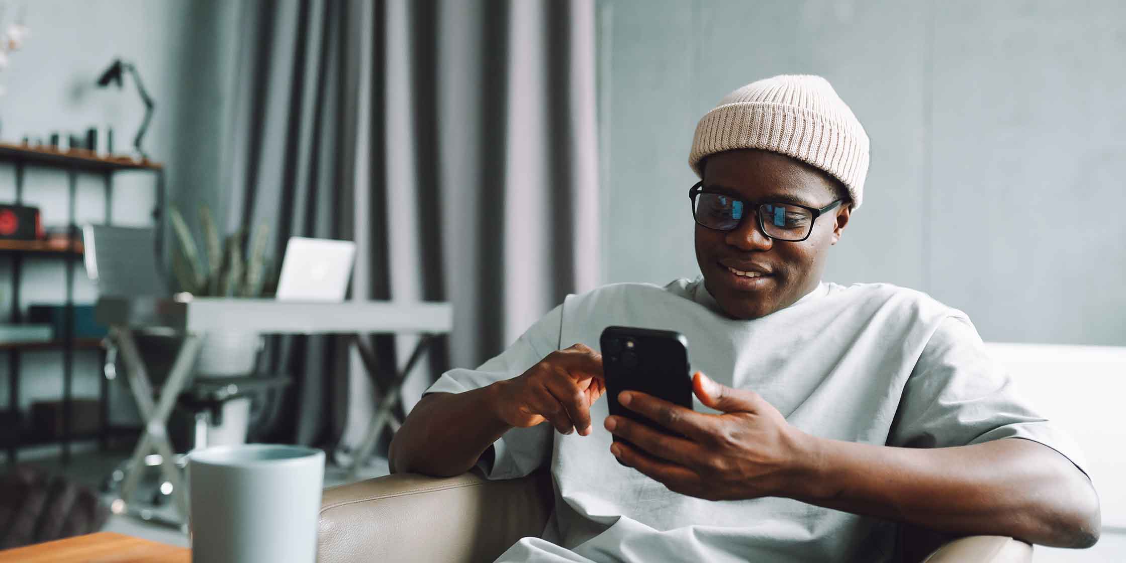 A smiling person wearing glasses and a watch cap looking at their smartphone.
