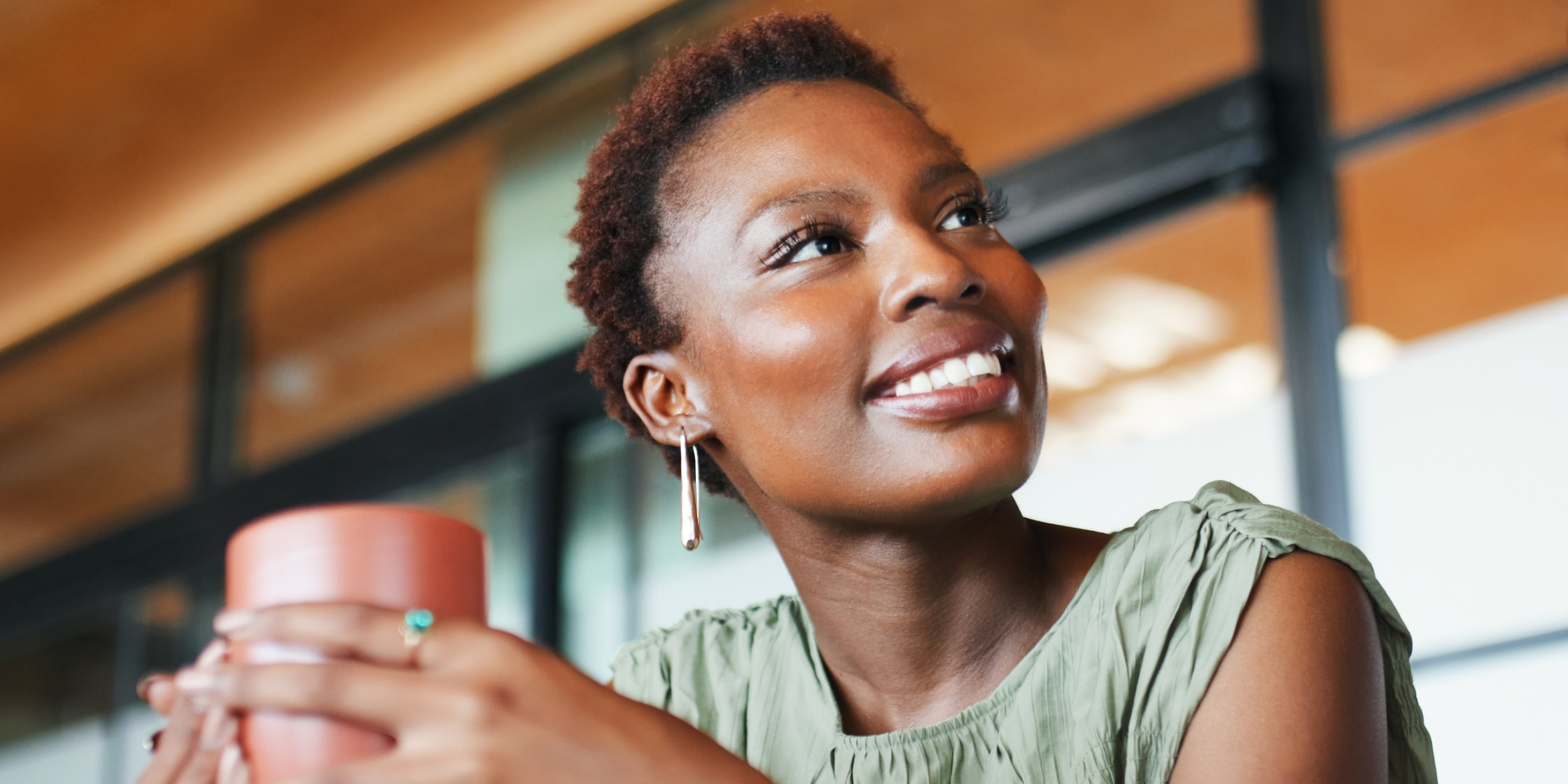 A smiling person sits in a coffee shop holding a mug of coffee.