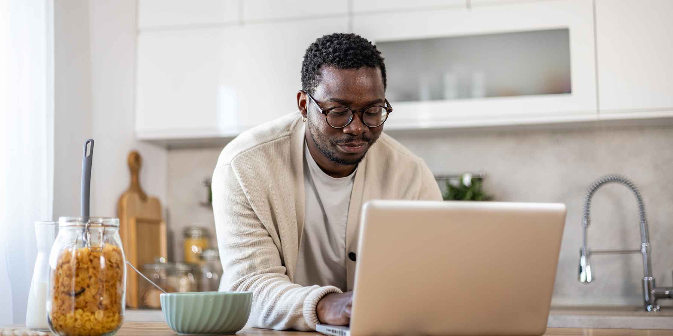 A smiling person in glasses looking at their laptop while standing at their kitchen counter.