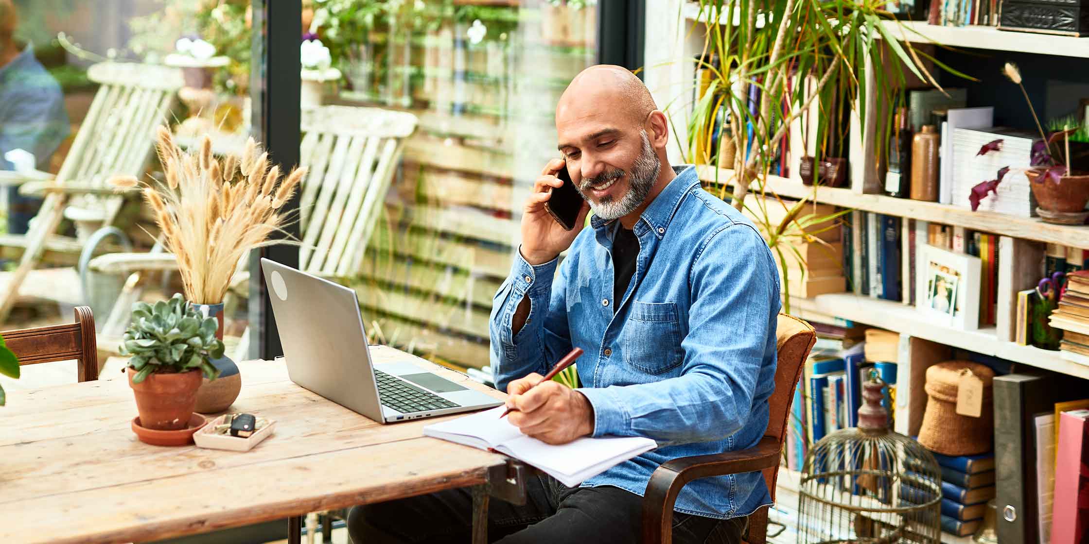 A person talking on the phone while taking notes and sitting in front of their laptop.