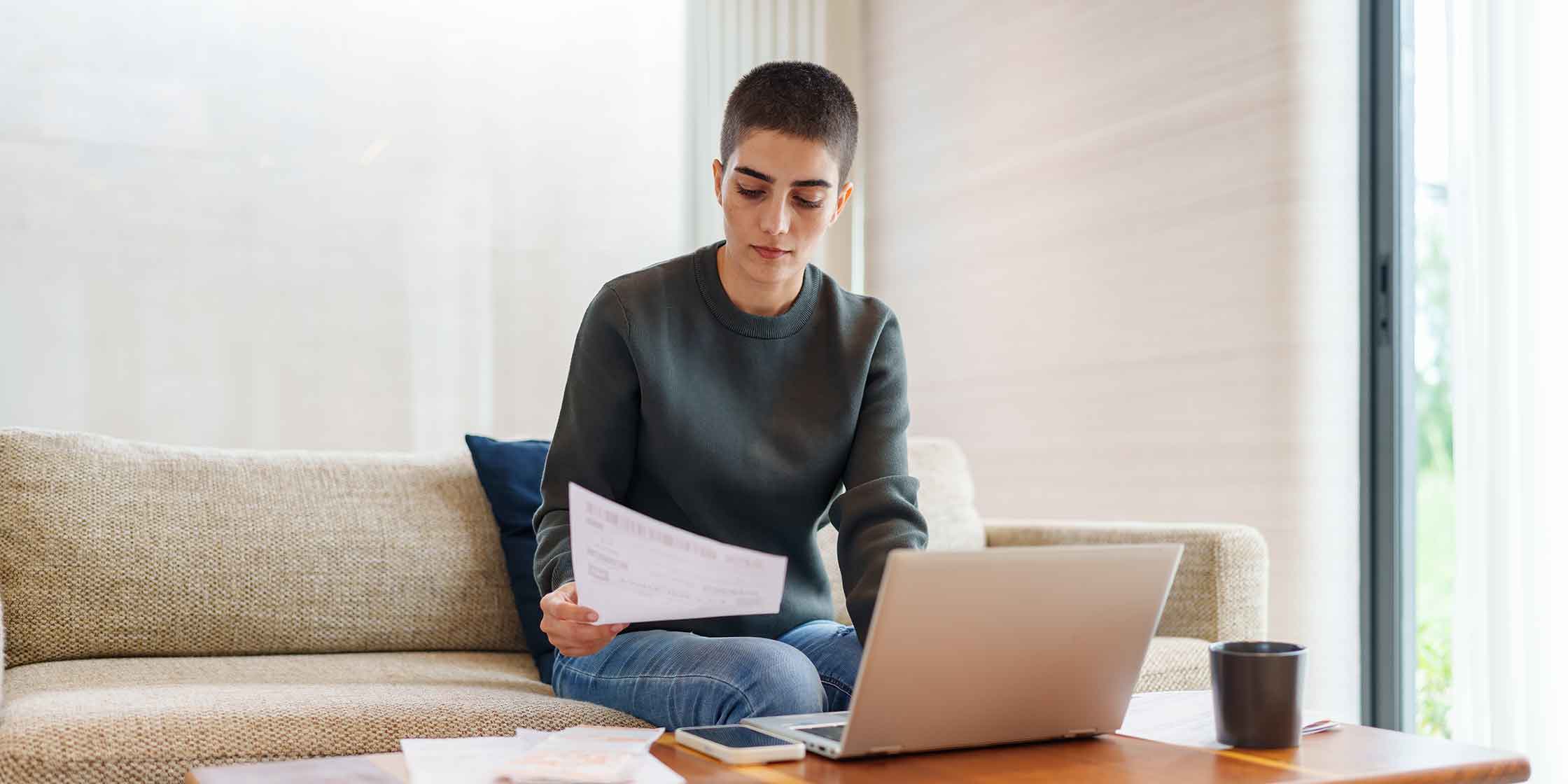 A person sits on a couch reading a document. An open laptop sits on a table in front of them.