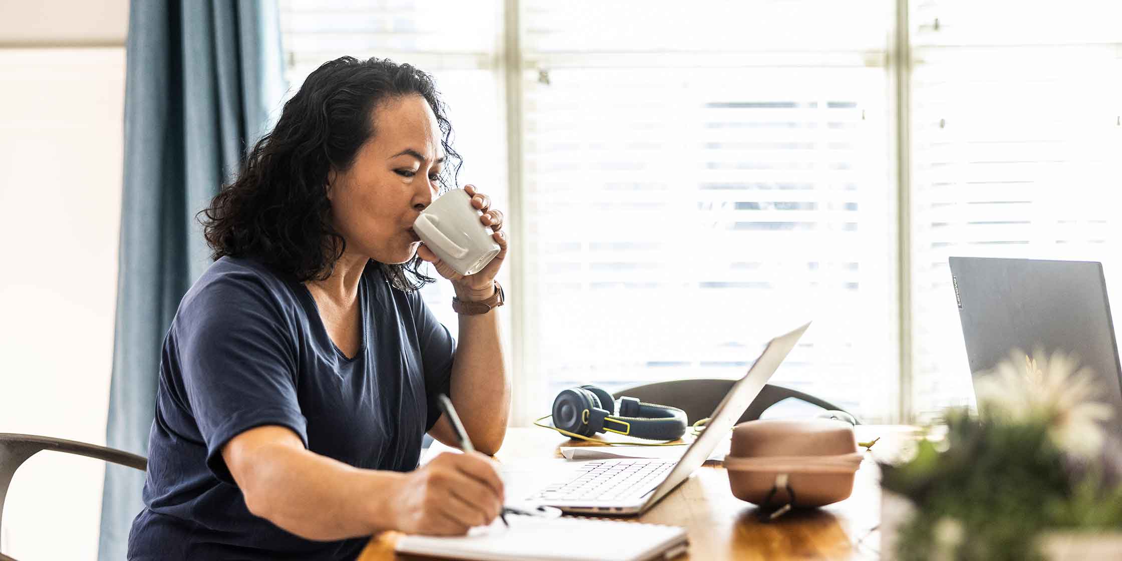 A woman sips from a mug while taking notes with a pen and paper in a bright home office.