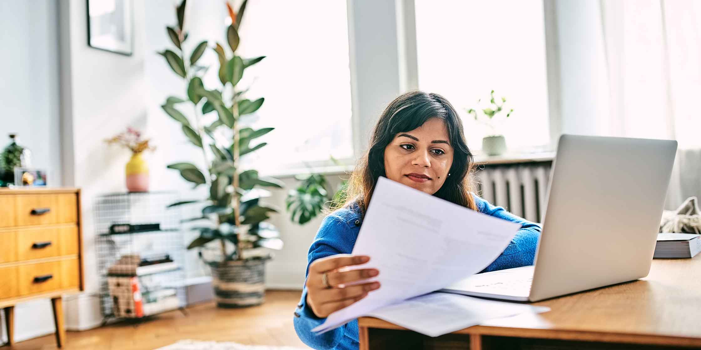 A person is in their home reading a paper document while sitting in front of an open laptop.