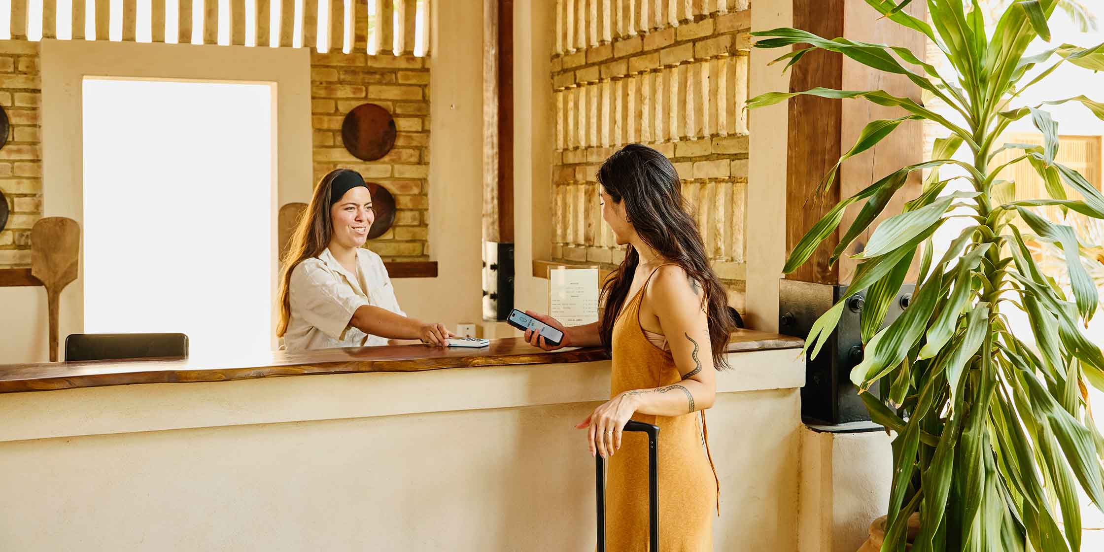 A woman holding a smartphone checks in at a hotel reception desk with a smiling staff member.