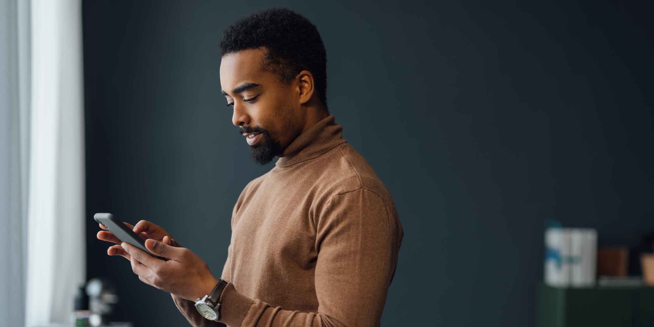 A man wearing a brown turtleneck thoughtfully uses his smartphone indoors against a dark backdrop.