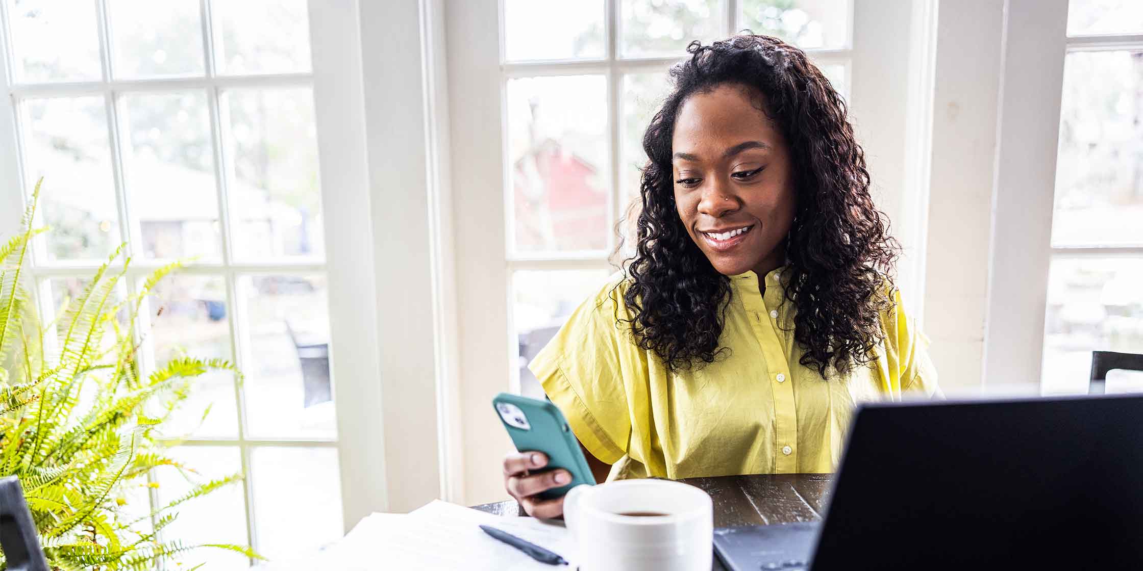 A woman smiles while looking at her phone and sitting in front of a laptop and a hot drink.