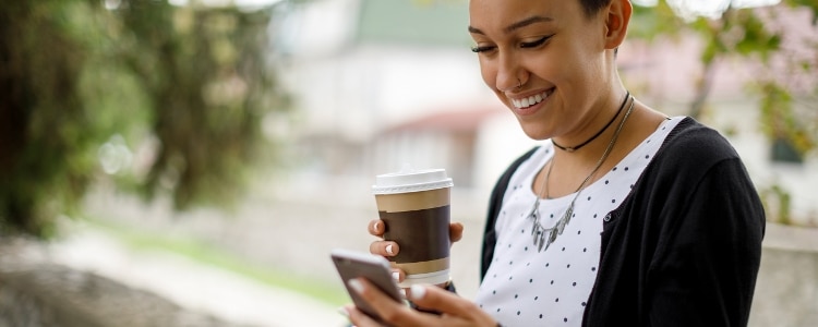 A woman holding a coffee cup smiles and looks at her phone.