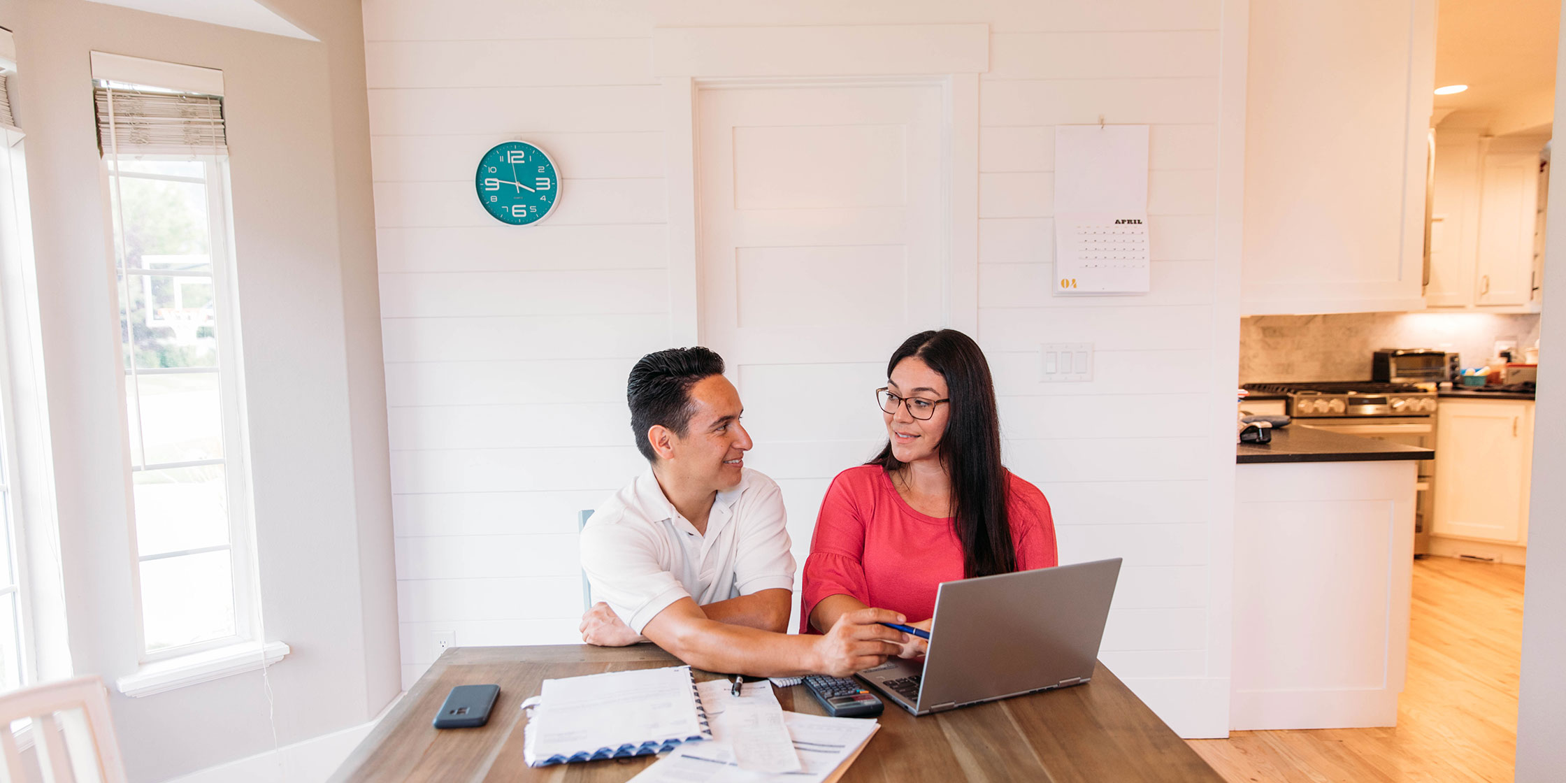 A smiling couple sitting at their kitchen table while looking at their laptop.