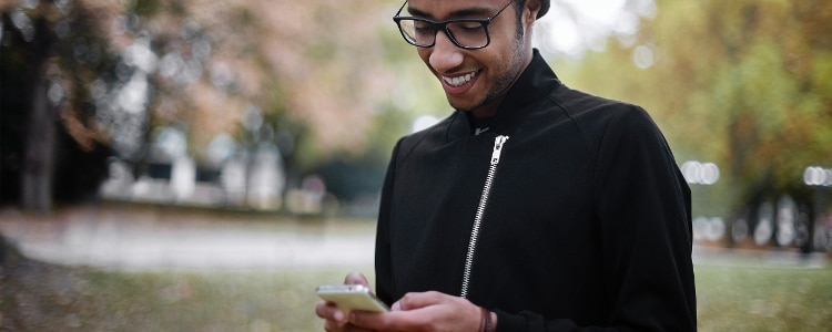 A man wearing a black jacket and glasses walks in the park looking at his phone.