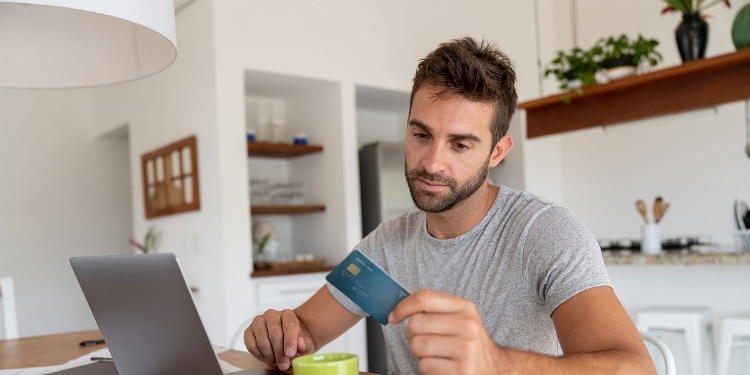 A person looking at their credit card while sitting at their kitchen table with their laptop and a cup of coffee.
