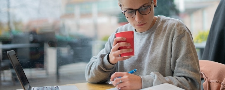 A person wearing glasses sits at their desk while drinking coffee and researching student loans.