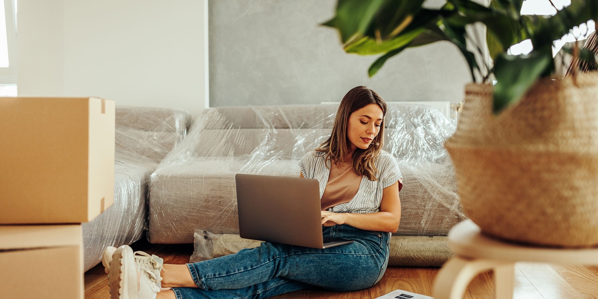 Someone sits on the floor and uses their laptop, surrounded by moving boxes and a couch wrapped in plastic.