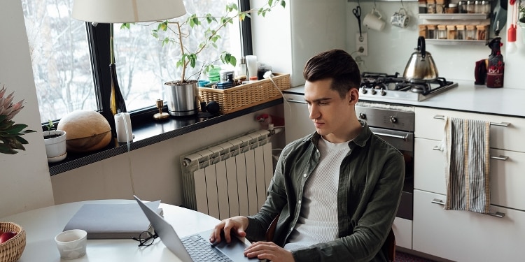 A person sits at a kitchen table with his laptop researching personal loans to pay off credit card debt.