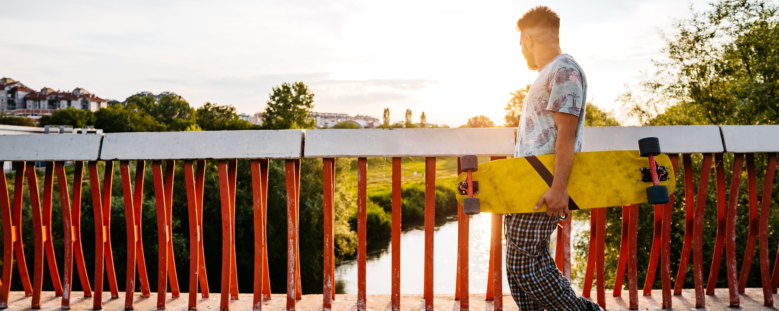 A person holding a longboard walks across a bridge and looks into the sunset in the distance.