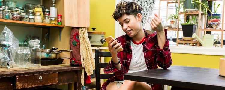 A person wearing a flannel shirt sits at their kitchen table, looking at their mobile phone.