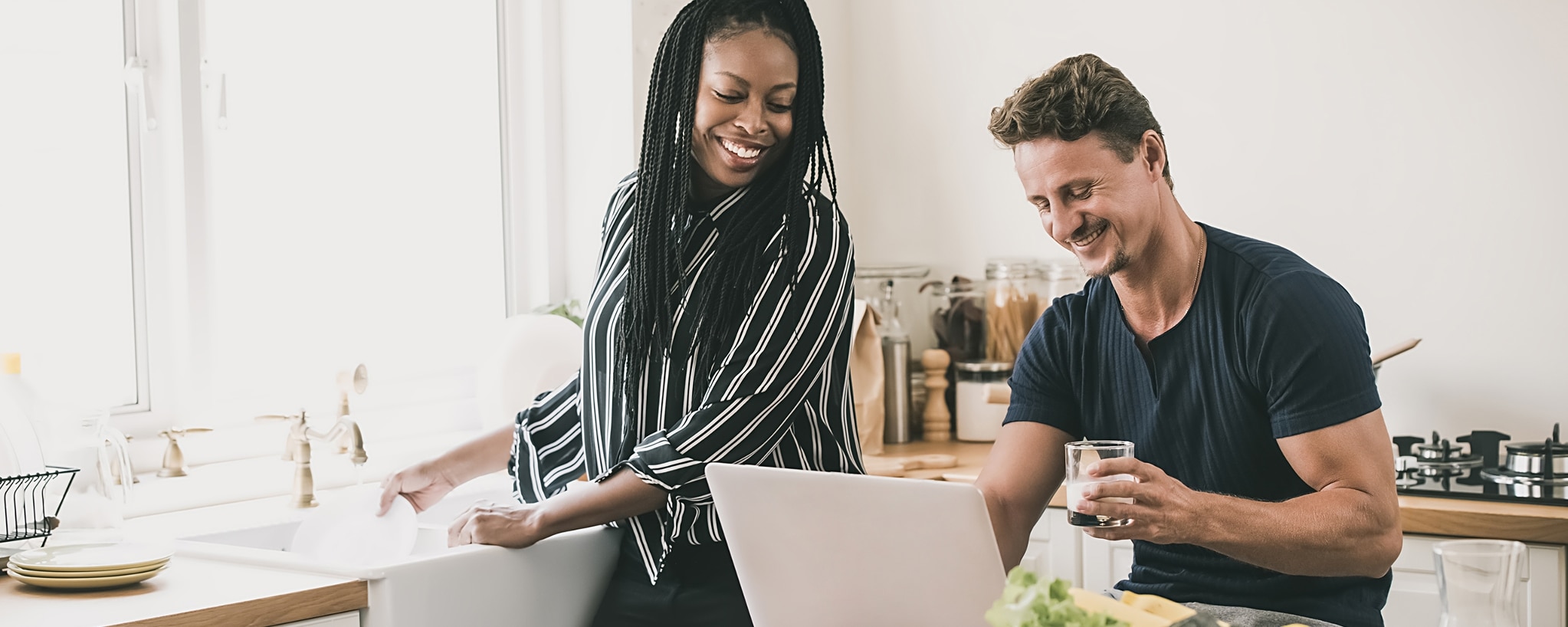Two people in a kitchen look at a laptop computer while one also washes dishes.