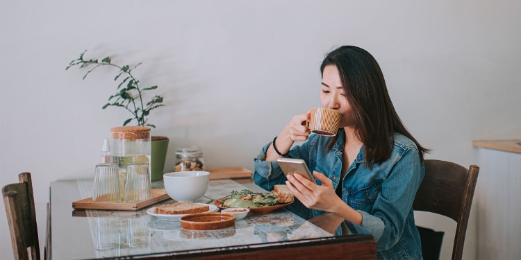 A person checks their FICO score on their phone while sitting at a table and drinking from a mug.
