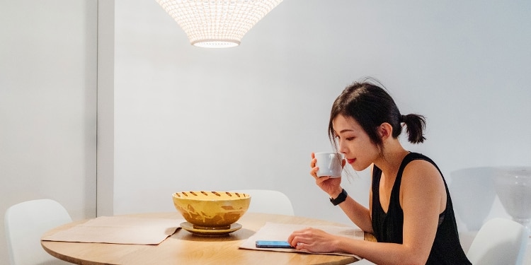 A person sits at their kitchen table while drinking coffee and using their phone.