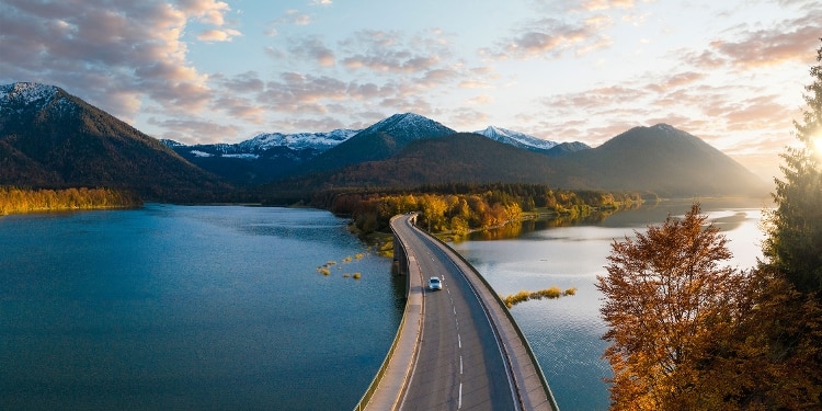 An aerial view of a person driving a rental car over a bridge and away from mountains.