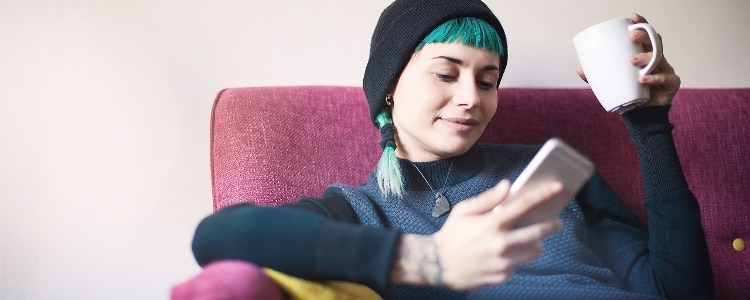 A person sits in a chair and looks at their phone while holding a mug.