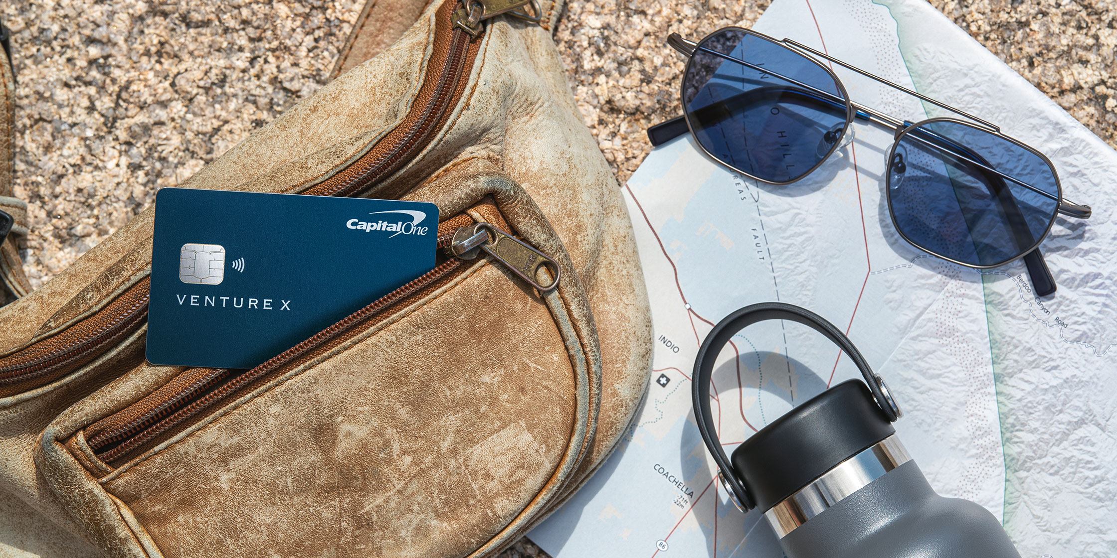A bag with a Capital One Venture X card in a zippered pocket rests on a rock with a map, a water bottle and a pair of sunglasses.