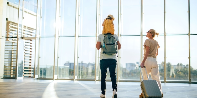 Two parents walk through an airport. One holds their child on their shoulders while the other pulls a rolling suitcase.