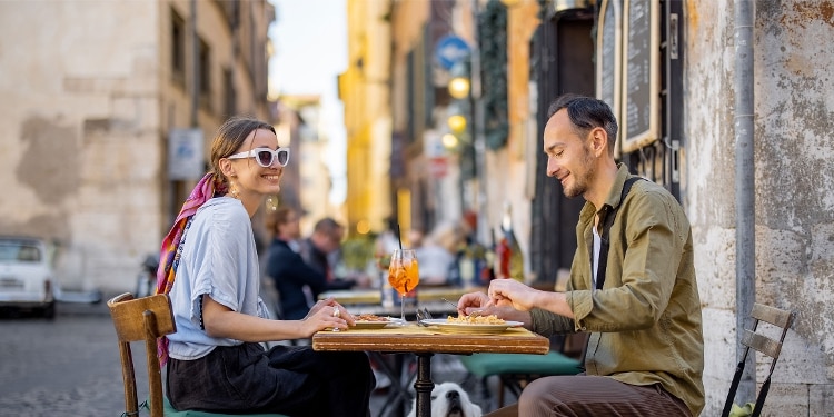 A smiling couple enjoying a meal and drinks outdoors in a European city.
