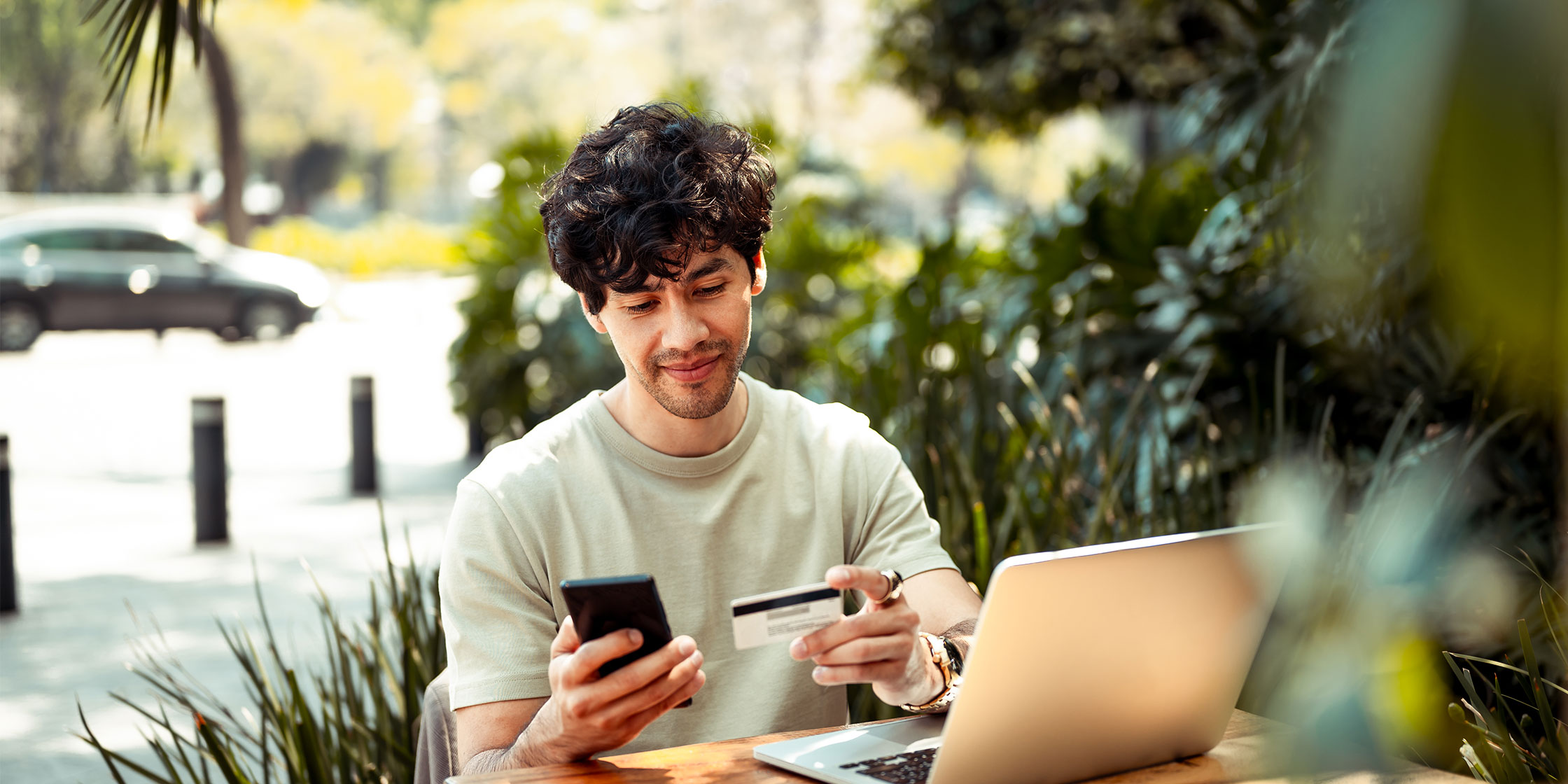 A young person sits at a table outside, holding a smartphone and looking at a credit card.