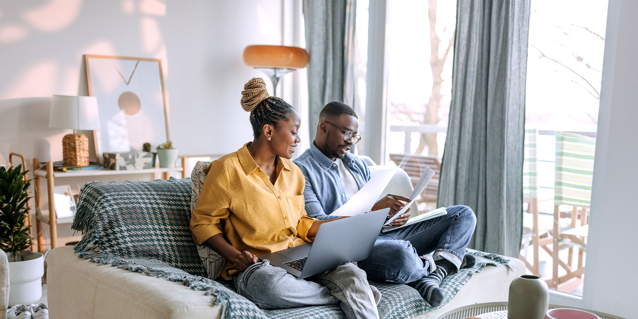 Two people sit on a couch while one uses a laptop and the other holds some documents.