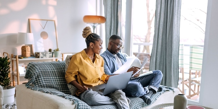 Two people sit on a couch while one uses a laptop and the other holds some documents.