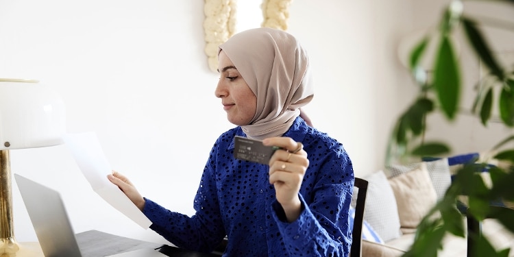 A woman sitting at a desk holds a credit card and looks at her credit card statement.
