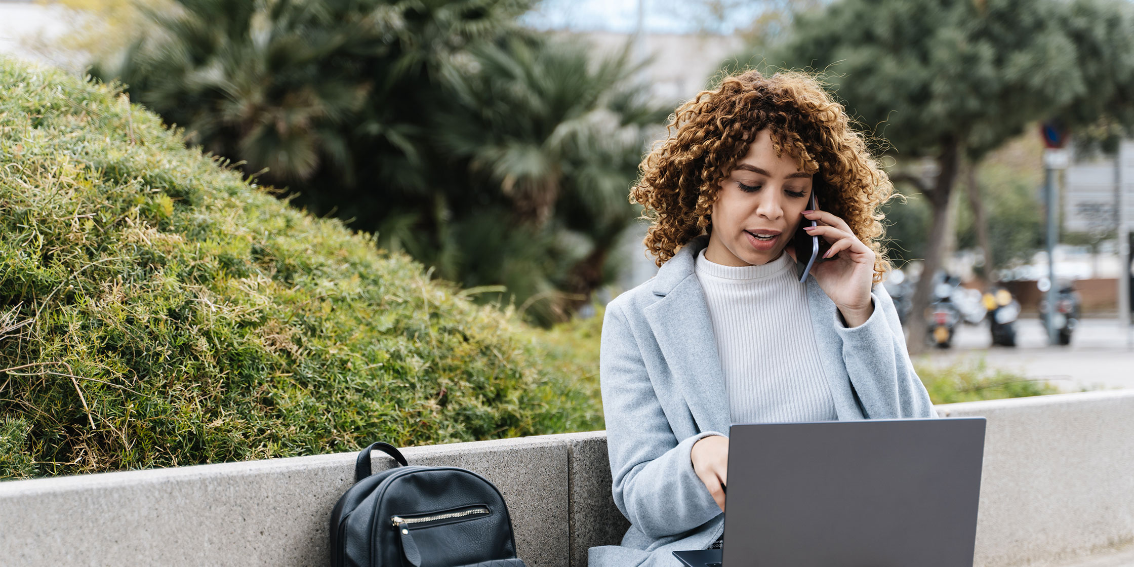 A person talking on their cellphone while sitting outside and looking at their laptop.