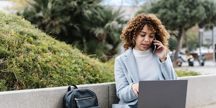 A person talking on their cellphone while sitting outside and looking at their laptop.